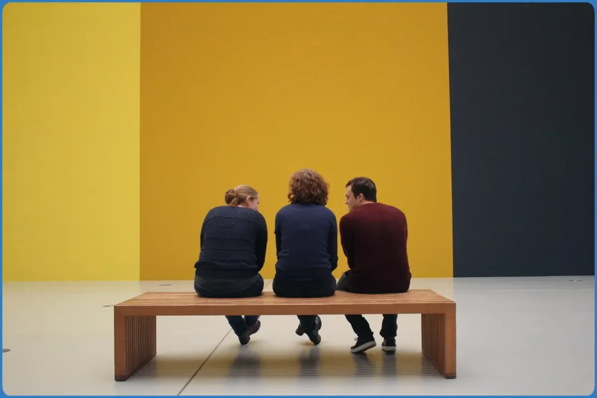 Three people sitting on a bench facing a yellow wall.