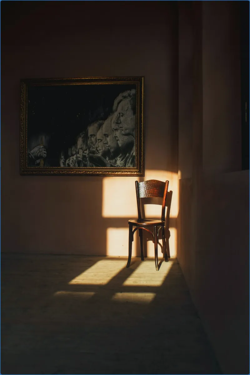 Chair in sunlight next to a framed painting on a wall.